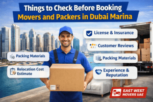 Professional movers holding a moving box in front of the skyline of Dubai Marina with a checklist showing things to check before booking movers and packers in Dubai by East West Movers UAE.